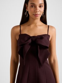 Temno bordo obleka z veliko pentljo na prsih, tankimi naramnicami in gubanimi detajli. Tkanina z rahlo teksturo in gladkim zaključkom.