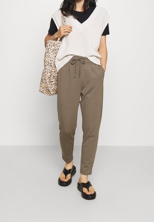 Person trägt braune Kordelhose, schwarzes T-Shirt, cremefarbene Strickweste, schwarze Sandalen und hält eine Schultertasche mit Blumenmuster.