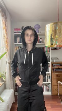 Sweatshirt preto com fecho, bolsos frontais, cordões e bainhaCanalizada. Modelo está de pé numa sala com estantes e uma lâmpada decorativa.