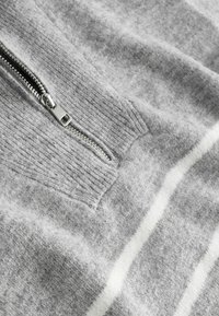 Grijze trui met ritssluiting en een gestructureerde stof, voorzien van een aansluitende kraag, een zilveren rits en witte horizontale strepen over de borst.