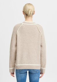 Beige trui van zachte stof met witte accenten. Heeft een geribbelde zoom, ronde halslijn en een ontspannen pasvorm. De achterkant toont raglanmouwen.