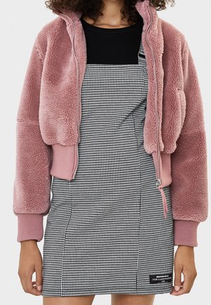 Woman wearing a black top, houndstooth patterned pinafore dress, and a cropped dusty pink fleece jacket with ribbed cuffs and zipper details.