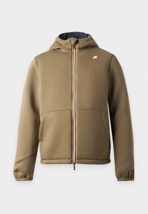Jeune homme debout portant un sweat à capuche zippé vert olive, un polo noir, un jean bleu clair et des baskets blanches avec des rayures noires.