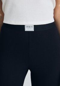 Ribbed black leggings with a wide elastic waistband. Features a "GUESS" label stitched on the front. Smooth and fitted design.
