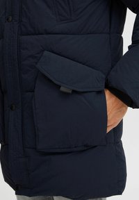 Chaqueta de plumas azul marino con un gran bolsillo frontal, cierres de botones y puños acanalados. La textura parece suave con un acabado ligeramente brillante.