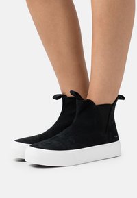 Svarta mocka höga sneakers med elastiska sidopaneler och vita gummisålar, med rundade tår och textildetaljer vid ankeln.