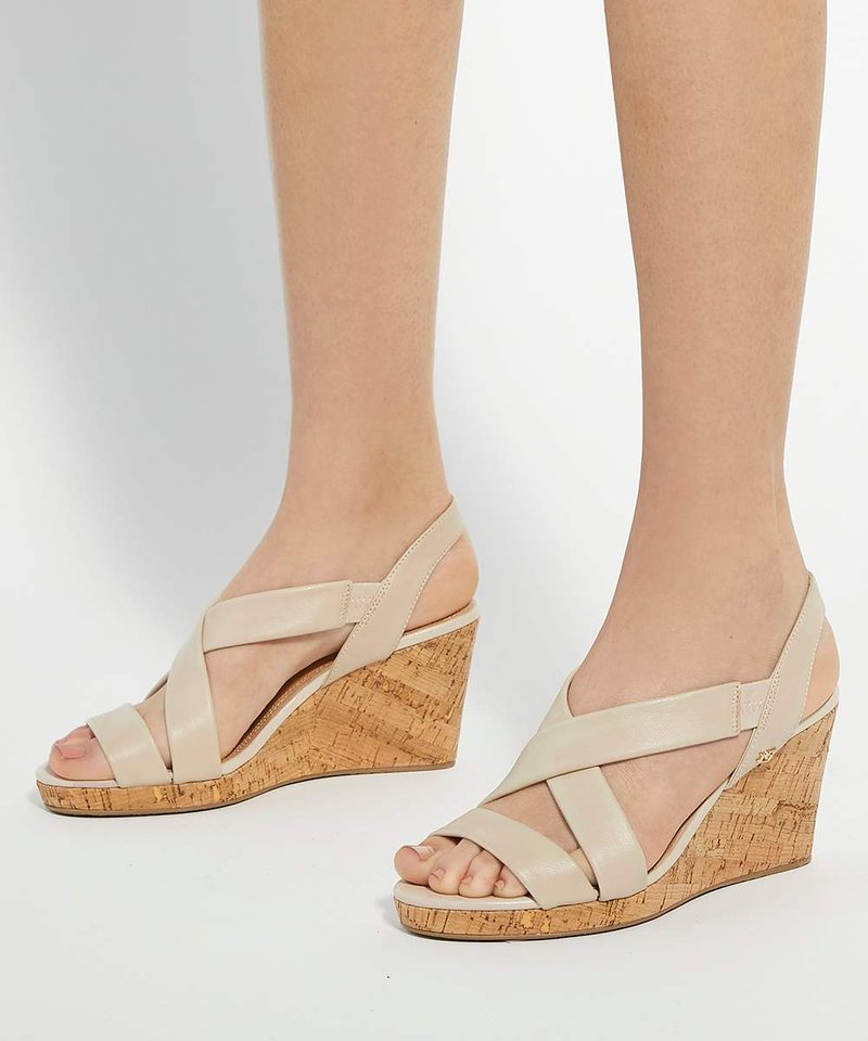 Pieds portant des sandales compensées en cuir beige avec talons en liège et brides croisées sur les orteils et autour des chevilles sur un fond blanc uni.