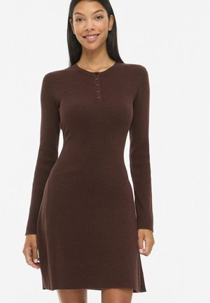 Femme souriante portant une robe marron en maille côtelée ajustée à manches longues avec des boutons au niveau de l'encolure sur un fond blanc uni.
