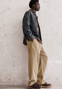 Džínová bunda na hnedom tričku; béžové kordové široké nohavice; hnedé topánky na šnurovanie s hrubou podrážkou; subjekt sa pózuje proti neutrálnej stene.