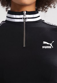 Detailní záběr černého trička Puma s bílým logem, se zipem s kulatým táhlem a černobílým proužkovaným vysokým límcem.