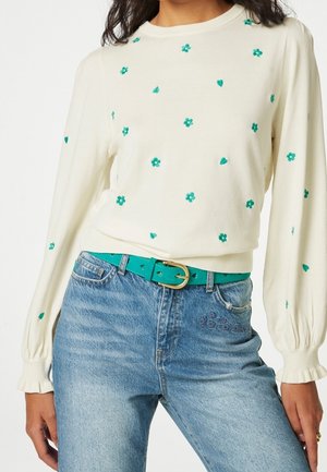 Maglione crema con ricami di fiori turchesi e cuori, abbinato a una cintura turchese coordinata, indossato con jeans blu chiaro.