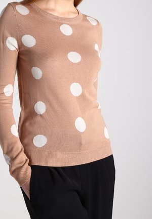 Béžový svetr s dlouhým rukávem s velkými bílými puntíky, nošený s černými kalhotami, ruce v kapsách, na šedém pozadí.