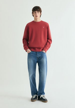 Młody mężczyzna stojący z rękami w kieszeniach, ubrany w czerwony sweter, niebieskie dżinsy i czarne trampki, na tle białej, gładkiej ściany.