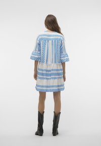 Robe blanche avec des motifs géométriques bleus, manches courtes et design à étages. Assortie à des bottes noires jusqu'aux genoux.