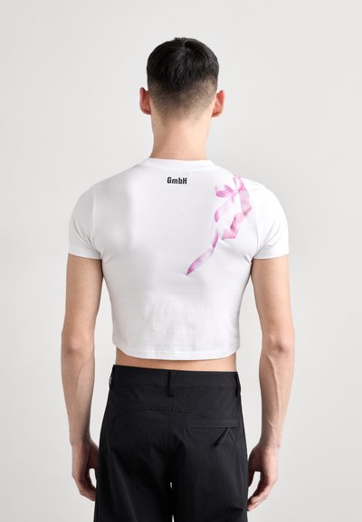 T-shirt blanc court avec un graphique de ruban rose au dos et le logo "GmbH" en noir au-dessus. Tissu lisse et design ajusté.