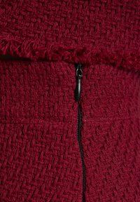 Rode gestructureerde stof met een geweven patroon, voorzien van een zwarte rits aan de naad en een franjesrand aan de bovenkant.