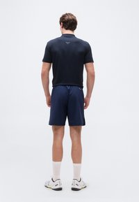 Άνδρας που φοράει μπλε ρουά πόλο μπλούζα, μπλε ρουά αθλητικό σορτς, λευκές κάλτσες και λευκά αθλητικά παπούτσια, στέκεται με την πλάτη προς τα εμπρός σε απλό φόντο.