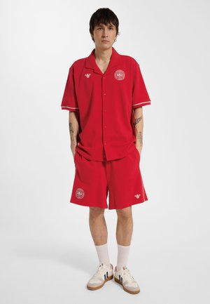 Junger Mann trägt ein rotes kurzärmeliges Hemd mit Knöpfen und passende Shorts mit weißen Logos, weiße Socken und weiße Sneakers vor schlichtem Hintergrund.