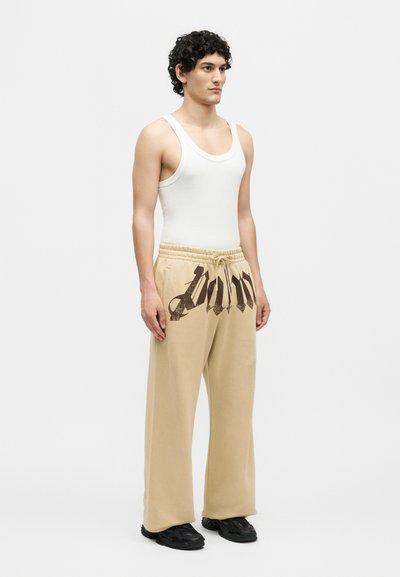 Νέος άνδρας στέκεται φορώντας λευκή φανέλα, μπεζ φόρμες με σκοτεινό στιλιζαρισμένο κείμενο στη μέση και μαύρα παπούτσια.