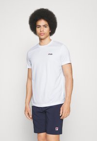 Camiseta atlética blanca de manga corta con un logo en el pecho, combinada con pantalones cortos azul marino que tienen un parche con logo. Textura suave.