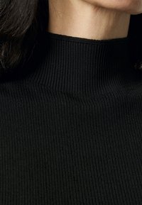 Musta ribatturjauhe, jossa on korkea kaulus, pehmeä koostumus ja istuva siluetti, korostaen materiaalin venyvyyttä ja kerroksellisuutta.