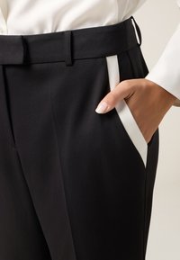 Zwarte broek van gladde stof, met witte zijzakken en een smalle tailleband. Er zit een hand in een zak ter schaal.