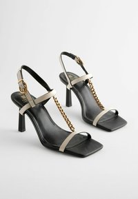 Schwarze High-Heel-Sandaletten mit quadratischen Zehen, ausgestattet mit beigen Riemen und goldenen Kettenakzenten. Aus Leder gefertigt, mit einem eleganten, modernen Design.