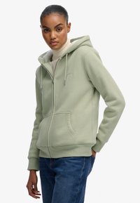 Vrouw in een lichtgroene hoodie met ritssluiting en capuchon, met voorzakken, gecombineerd met blauwe jeans en een crèmekleurige coltrui.