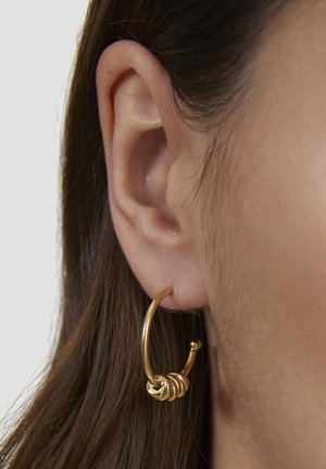 Close-up van een linker oor met een gouden oorring en meerdere kleine gouden ringen die onderaan bij elkaar zijn gegroepeerd.