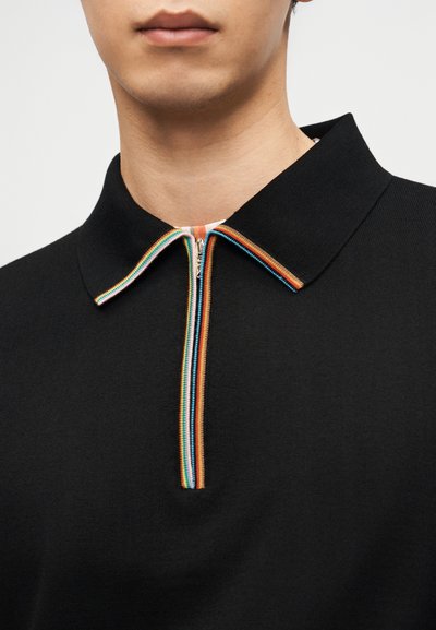 Άνδρας που φοράει μαύρο πόλο πουκάμισο με πολύχρωμη ρίγα στο φερμουάρ και το περίγραμμα του γιακά, εμφανιζόμενος από τον λαιμό μέχρι το στήθος.