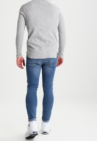 Maglietta grigia a maniche lunghe con cucitura posteriore, abbinata a jeans blu e sneakers bianche. I jeans hanno tasche tradizionali e una vestibilità slim.