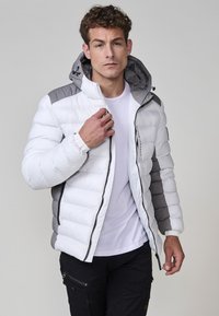 Giacca imbottita bianca con accenni grigi, caratterizzata da una chiusura a zip, materiale sintetico, cappuccio removibile e polsini elasticizzati.