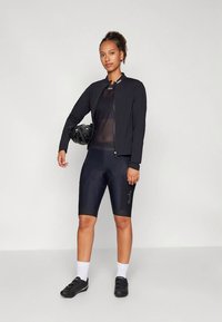 Nainen mustassa pyöräilytakissa, verkkopaidassa, topatuissa pyöräilyshortseissa, valkoisissa sukissa ja mustissa pyöräilykengissä, pitää mustaa kypärää sivullaan.