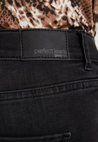 Jean en denim noir avec un tissu texturé. Présente une étiquette rectangulaire noire indiquant "jeans parfaits" et "ginatricot" en lettres blanches.