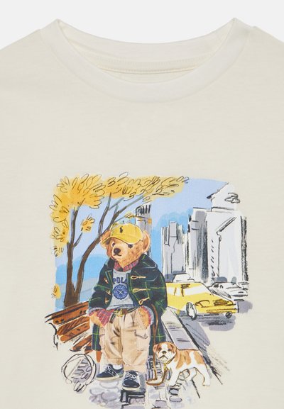 Krämfärgad t-shirt med en tecknad björn i grön jacka, gul keps och beige byxor, som går med en hund med en stadsbackgrund.