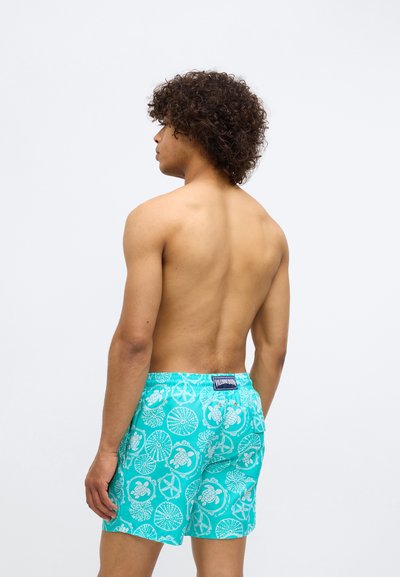 Turkizne kopalke z belimi vzorci, ki prikazujejo morsko tematiko, vključno s želvami in pomorskimi simboli. Tkanina je lahka in gladka.