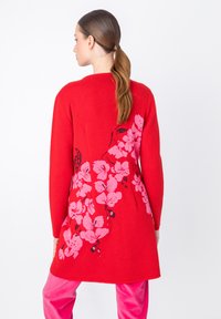Červený pletený kardigán s dlhými rukávmi, na zadnej strane s ružovým kvetinovým vzorom. Hladká textúra, dĺžka do polovice stehien, bez viditeľného kovania.