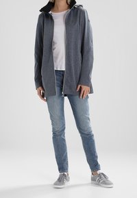 Šedá zipová bunda s vysokým límcem, s kontrastní černou kapucí. Nositá přes světle barevné tričko, kombinovaná s modrými džíny a šedými botami.