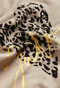 Tkanina s leopardím potiskem v béžové barvě s černými skvrnami. Vzorek doplněn zlatavým, kurzívním textem přes vzor. Měkká textura, zmačkaný vzhled.