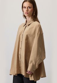 Lihtne oversize nuppudega särk, millel on terav krae, pikad varrukad ja tekstureeritud kangas, ning millel on lõhenenud hem ja esikoti detail.