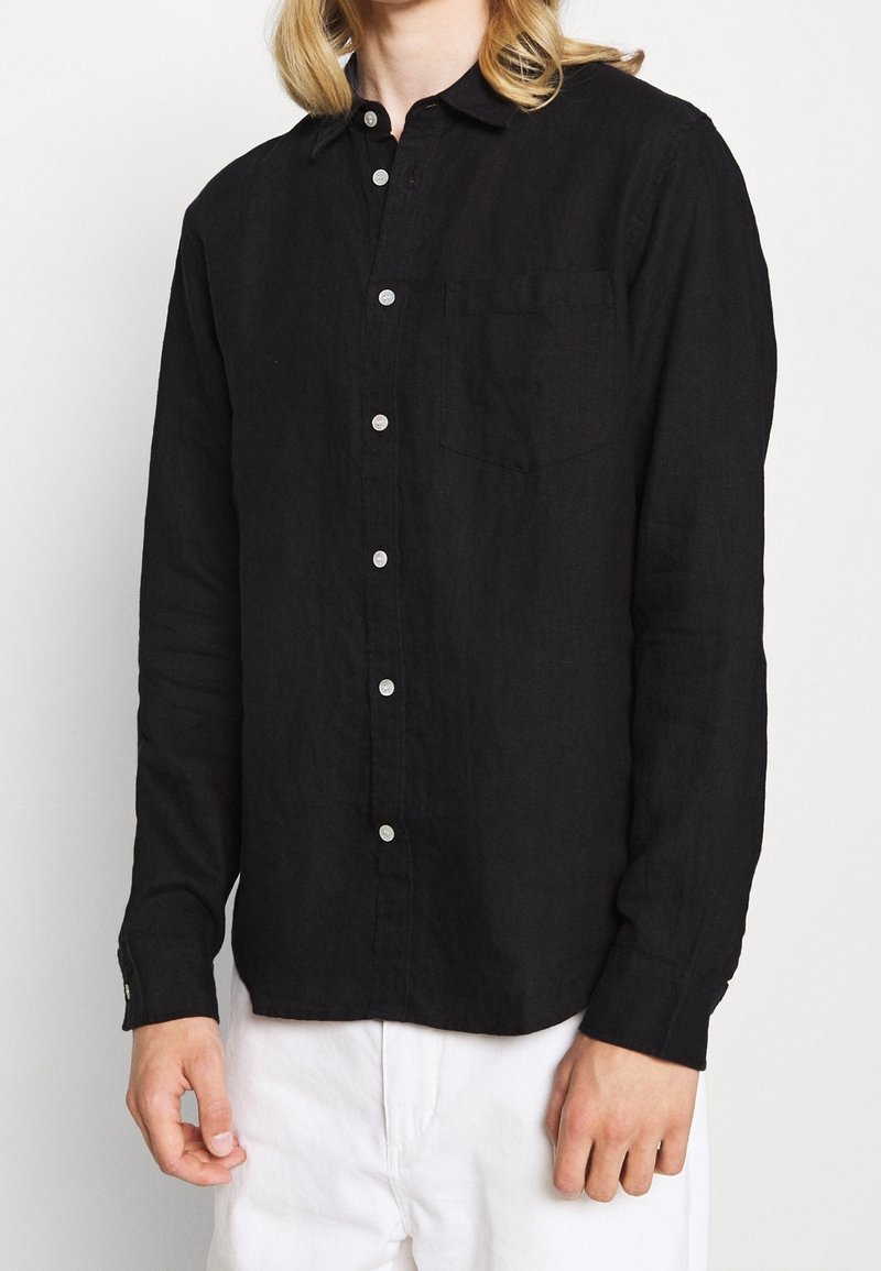 Personne portant une chemise noire à manches longues avec une poche poitrine et un pantalon blanc, debout devant un fond uni.