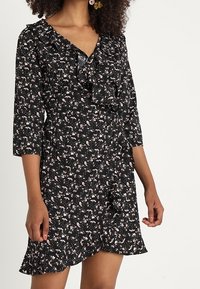 Musta kukkamekko, jossa on kolmanneksen hihat, röyhelökaulus ja epäsymmetrinen helma. Kangas sisältää pieniä vaaleanpunaisia ja valkoisia kukkakuoseja.