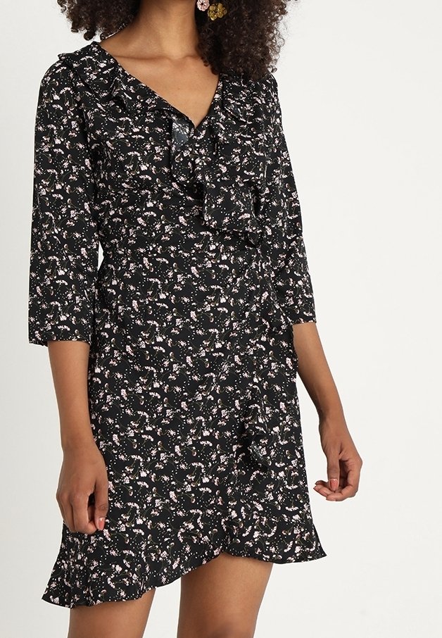 Musta kukkamekko, jossa on kolmanneksen hihat, röyhelökaulus ja epäsymmetrinen helma. Kangas sisältää pieniä vaaleanpunaisia ja valkoisia kukkakuoseja.