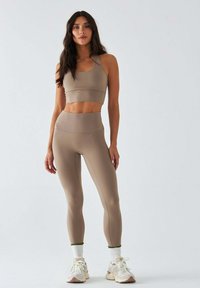 Beżowy sportowy biustonosz i pasujące legginsy z wysokim stanem, wykonane z elastycznego materiału, z subtelnymi szwami. Sneakersy z białymi i pastelowymi akcentami.