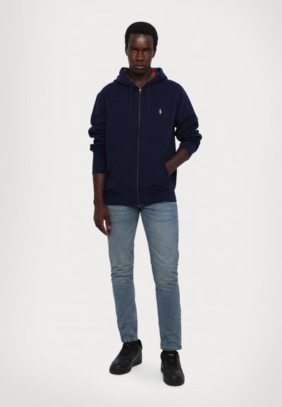 Veste à zip marine avec une poche kangourou et une capuche à cordon, assortie à un jean bleu clair et des baskets noires. Petit logo en accent sur la poitrine.