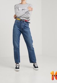 Lichtgrijs shirt met lange mouwen en logo, hoog getailleerde blauwe spijkerbroek, meerkleurige riem en zwarte sneakers met witte zolen.