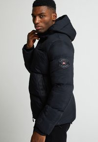 Melna pūkainā jaka ar rāvējslēdzēju, kapuci un ribotām piedurknēm. Uz kreisās piedurknes ir aplis ar apdruku. Gluda tekstūra, silts dizains.