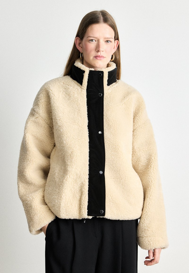 Μπεζ fleece ζακέτα με μαύρο καπιτονέ μπροστινό πάνελ, κλείσιμο με κουμπιά snap και ψηλό γιακά. Διαθέτει χαλαρή εφαρμογή και φαρδιά μανίκια.