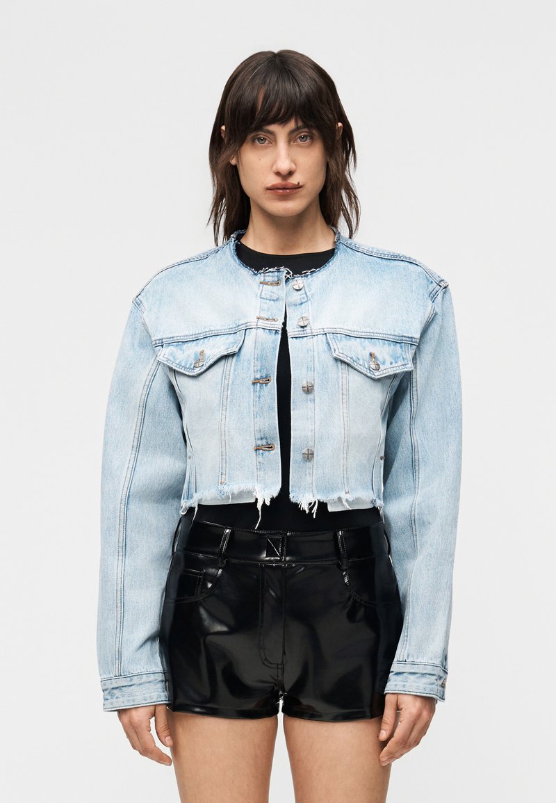 Νεαρός με σκούρα μαλλιά μέχρι τους ώμους, φορώντας ένα κοντό ελαφρύ τζιν σορτς και μαύρο λαμπερό σορτς, στέκεται μπροστά σε απλό φόντο.