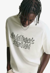 Krémové tričko s krátkým rukávem a černým vyšitým textem "laidback stories" na hrudi. Měkká látka, uvolněný střih, minimalistický design.